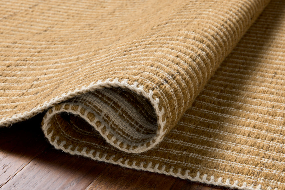 Elm wheat  beige rug on a wooden floor. Slightly rolled to show thickness. Edged in white cotton stitching 