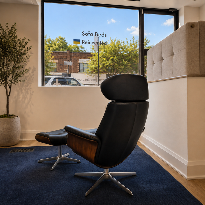 Timeless black leather recliner with a matching ottoman, is placed on a dark blue rug in a bright, contemporary room. Large windows behind the furniture show a street scene with trees and a sign reading 'Sofa Beds Reinvented', and a tufted beige sofa is visible in the background.