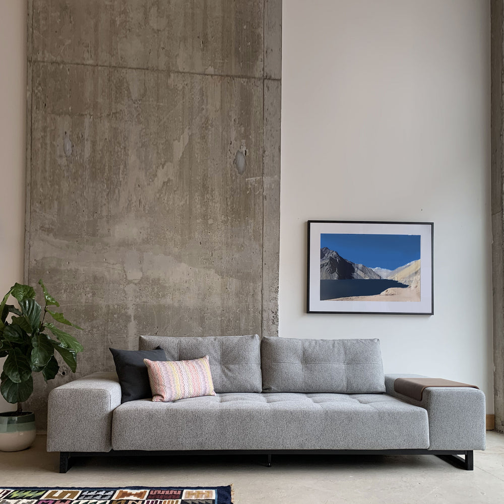A gray Soho Sofa Bed with ottoman in a minimalist interior setting, featuring a wall of concrete and a framed abstract picture above the sofa.